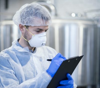 Technologist specialist with protective mask and hairnet holding clipboard checklist and writing production results.